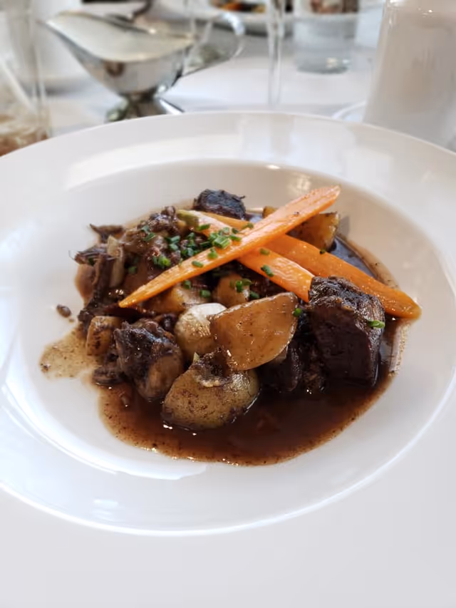 A white bowl filled with a stew containing chunks of beef, potatoes, carrots, and garnished with chopped chives, placed on a white table with blurred glassware and condiments in the background.