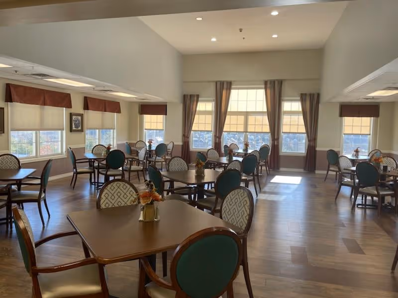 Bright dining room with multiple round tables and upholstered chairs arranged near large windows with shades and curtains.