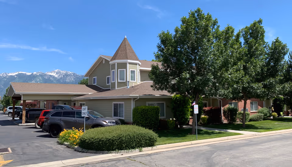 Exterior view of The Lodge at Jordan River facility showing a two-story building with a turret-like architectural feature, surrounded by trees, bushes, and a parking area with several cars. Mountains are visible in the background under a clear blue sky.