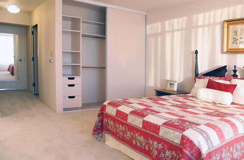 Bedroom with a red-and-white patchwork quilt on the bed, an open closet with shelves and drawers, a nightstand and a hallway with a mirror.