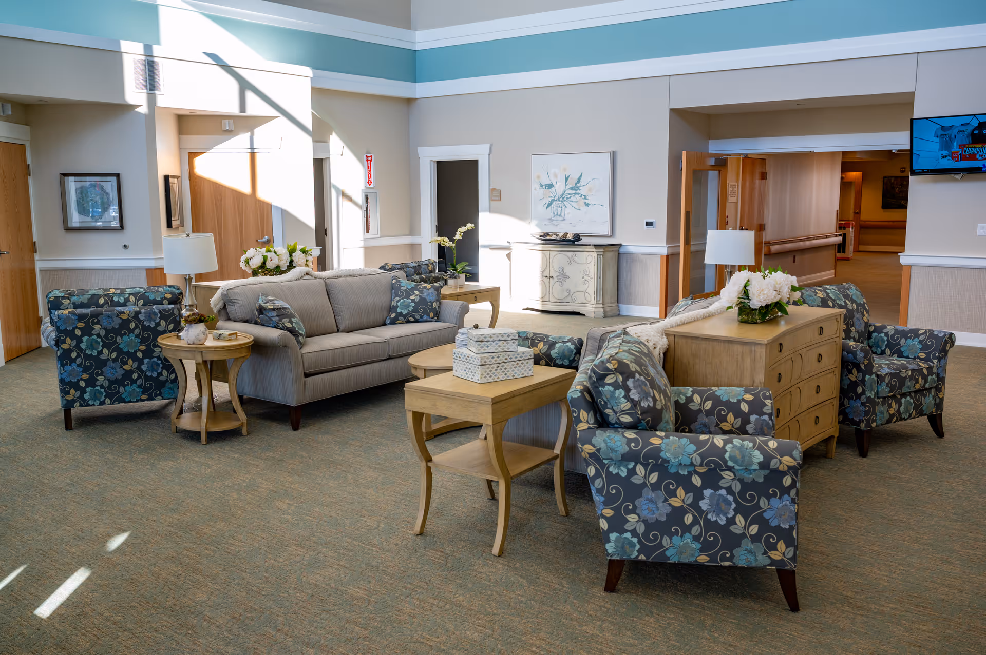 A bright and spacious living room area in a senior living facility with a beige sofa, four floral-patterned armchairs, wooden side tables, and a wooden chest with a flower arrangement on top. The room has carpeted flooring, light-colored walls, and a wall-mounted TV in the background.