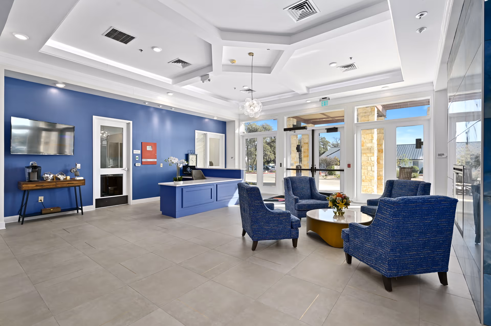 A bright and modern lobby area with a blue accent wall, a reception desk, four blue upholstered armchairs arranged around a round coffee table with a flower arrangement, large windows and glass doors letting in natural light, and a mounted flat-screen TV on the wall.