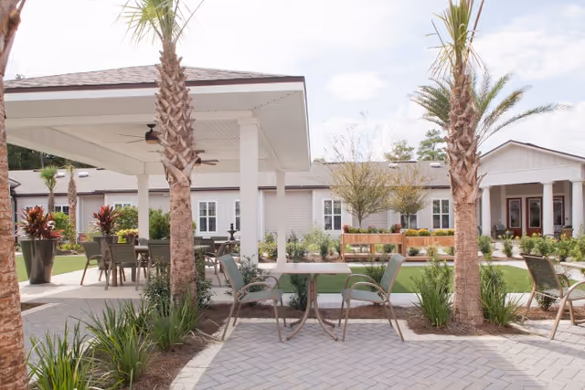Outdoor patio area at Ortega Gardens featuring a covered seating area with tables and chairs, palm trees, landscaped greenery, and a building with white siding and multiple windows in the background under a partly cloudy sky.