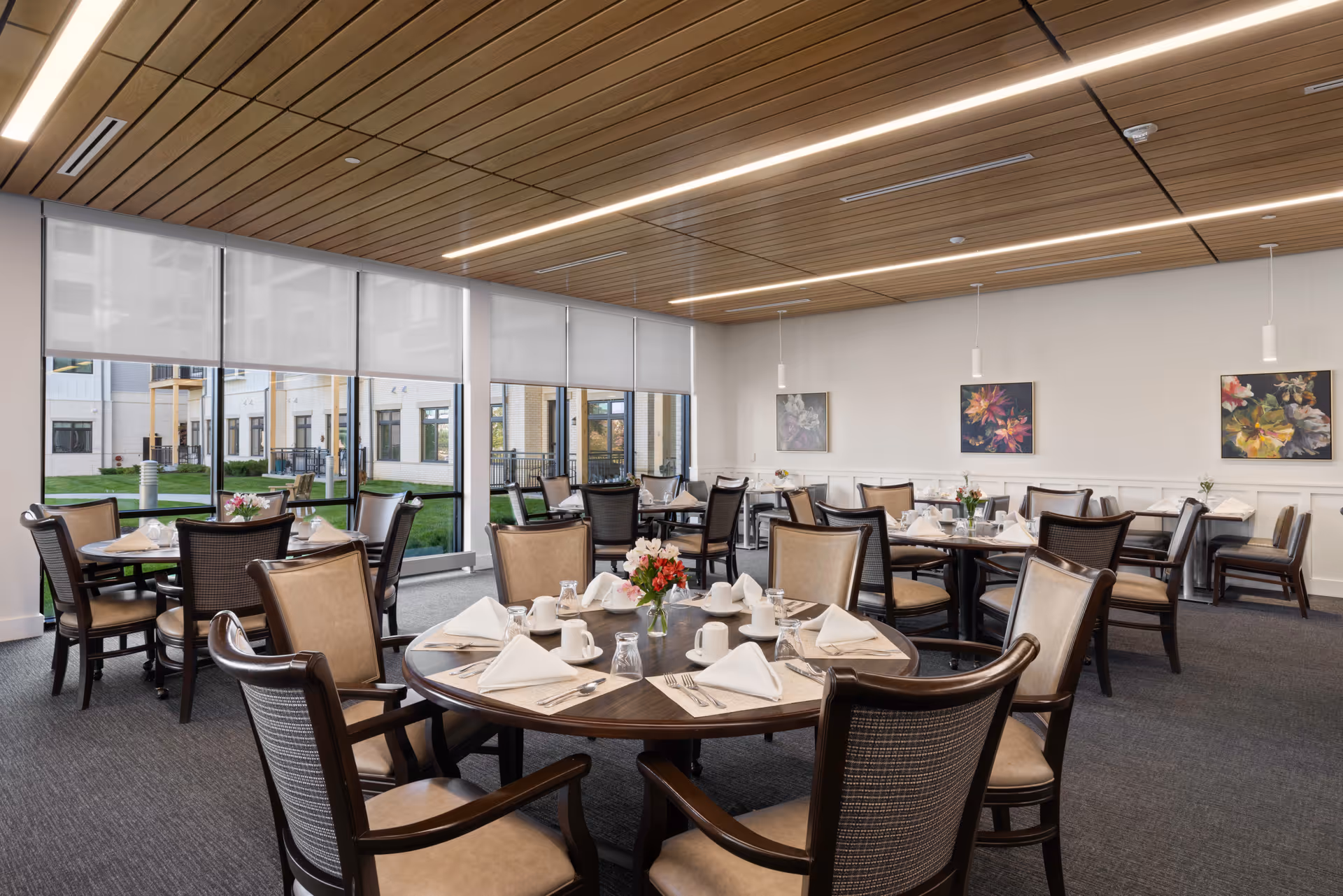 A bright and spacious dining room in a senior living facility with multiple round tables set for a meal. Each table has beige cushioned chairs, white napkins folded neatly, cups, glasses, and silverware. The room features large windows with roller shades, allowing natural light to fill the space. The ceiling has wooden paneling with modern linear lighting, and the walls are decorated with colorful floral artwork.
