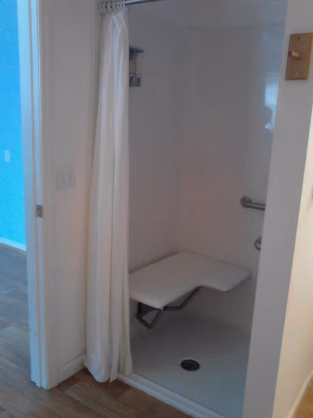 A small shower area with a white fold-down bench attached to the wall and a white shower curtain partially drawn to the side. The shower has a grab bar on the wall and a drain on the floor. The surrounding walls are white, and the floor outside the shower is wooden.
