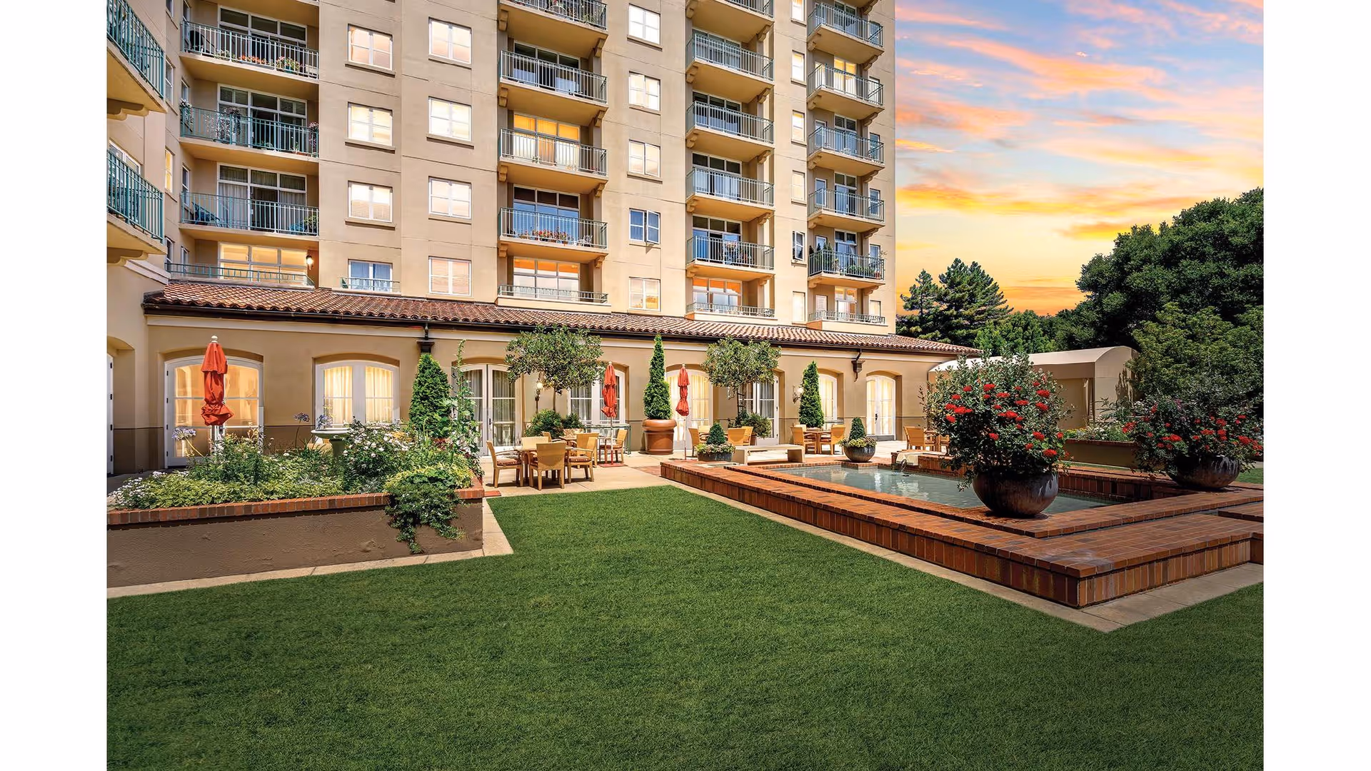 Outdoor courtyard with green lawn, raised rectangular fountain, patio tables, umbrellas and planters in front of a multi-story residential building at sunset.
