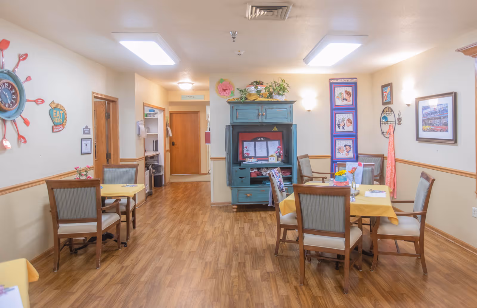 Small dining room with several tables and chairs, a decorative cabinet, and wall art in a senior living facility.