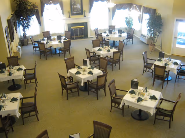 A dining room with multiple square tables covered with white tablecloths, each set with plates, cups, and green napkins. The room has large windows with curtains, a fireplace with a decorative screen, and several potted plants. Chairs are arranged around each table, and the room is well-lit with natural light.