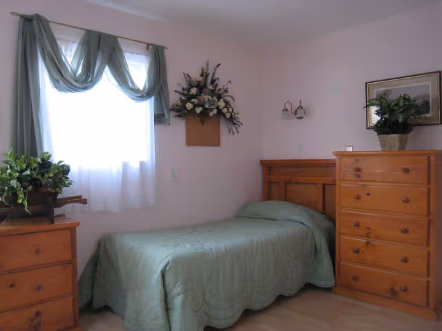 A small bedroom with a single bed covered in a green quilt. There is a wooden dresser with a potted plant on top to the right of the bed, and another wooden dresser with a plant on the left side near a window with sheer white curtains and green drapes. A floral wall decoration hangs above the bed.
