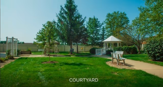 A sunny courtyard with green grass, trees, a white gazebo, a wooden bench, and a paved walkway. The area is fenced and has a swing set and garden features.