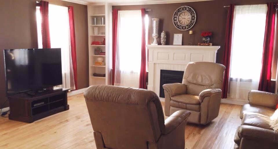 A cozy living room with three brown leather armchairs arranged around a white fireplace. The room has wooden flooring, a large flat-screen TV on a dark wooden stand, and three windows with white sheer curtains and red drapes. Decorative items including a large clock, vases, and framed pictures are placed on the fireplace mantel.