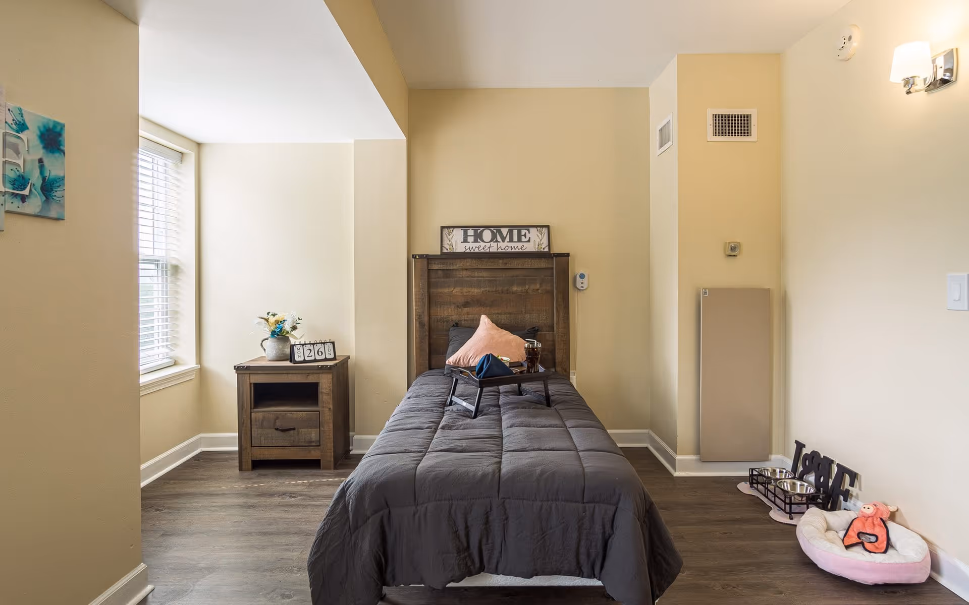 A cozy bedroom with a single bed covered in a dark gray comforter, a pink pillow, and a small tray with a cup and napkin on the bed. Behind the bed is a wooden headboard with a decorative sign that reads 'HOME sweet home'. To the left of the bed is a wooden nightstand with a vase of flowers and a calendar block showing the number 26. The room has beige walls, a window with white blinds, and wood flooring. On the right side near the wall is a pet bed with a pink toy and two pet bowls on a stand.