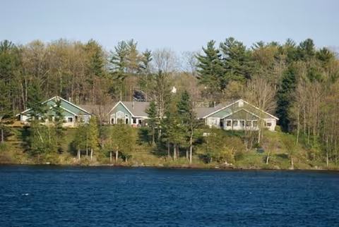 View of a group of buildings surrounded by trees on a hillside overlooking a large body of water under a clear sky.
