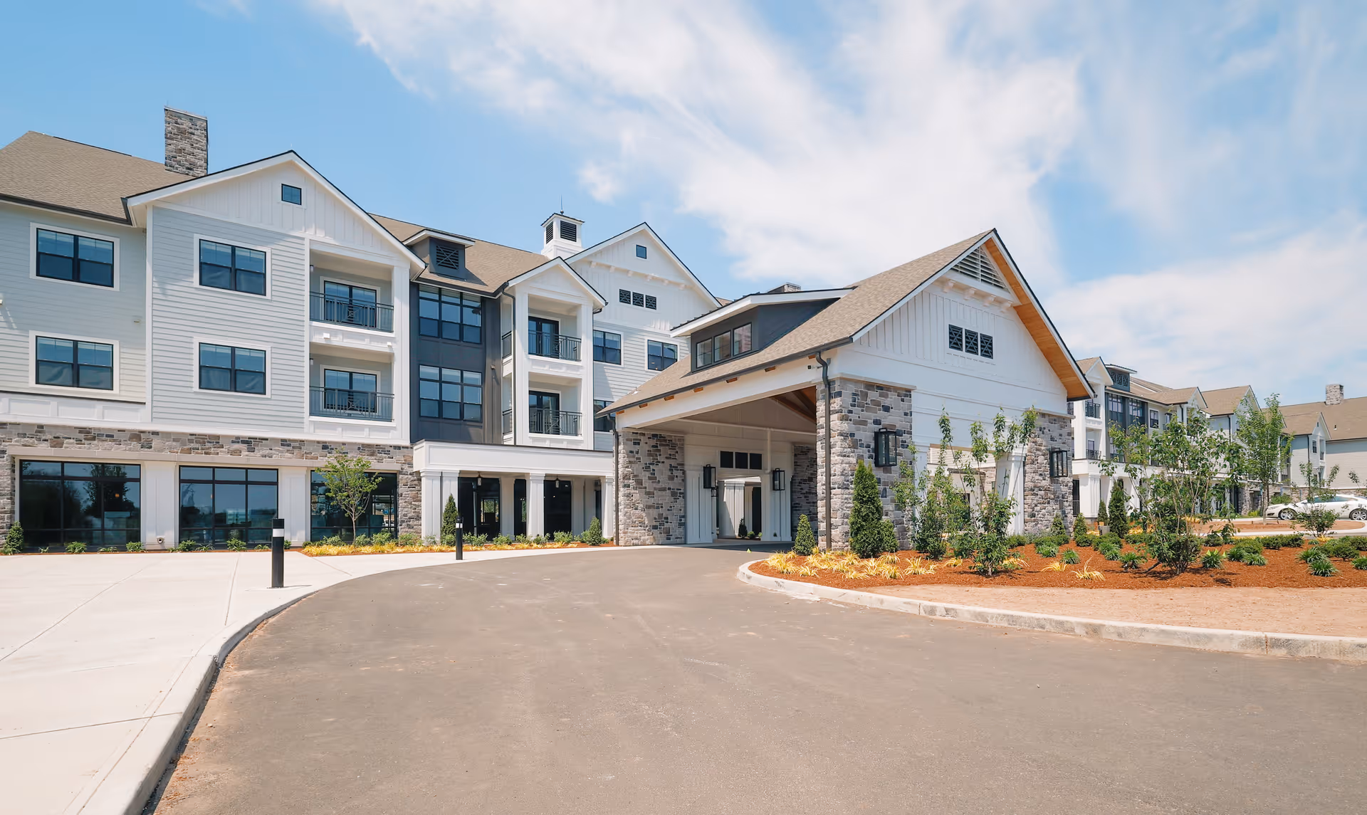 Front entrance of a multi-story senior living facility with a covered porte-cochere, balconies, and landscaped driveway under a blue sky.