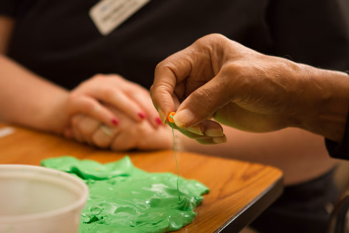 Close-up of an elderly person's hand stretching green slime or putty on a wooden table, with another person in the background holding their hands together.