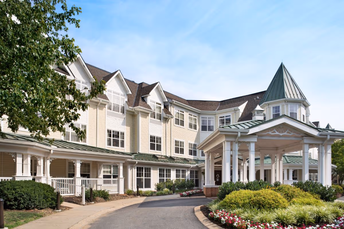 Exterior view of Sunrise of Lafayette Hill senior living facility showing a large, multi-story building with cream-colored siding and white trim. The building features multiple windows and a covered entrance with a green metal roof and a pointed turret. There are landscaped bushes and flowers along the driveway leading to the entrance under a clear blue sky.