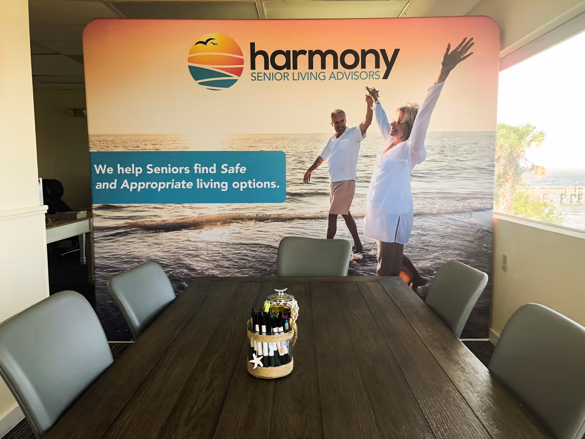 Conference room with a wooden table and six gray chairs. On the wall is a large banner for Harmony Senior Living Advisors featuring an image of a senior couple walking on a beach at sunset and the text 'We help Seniors find Safe and Appropriate living options.'