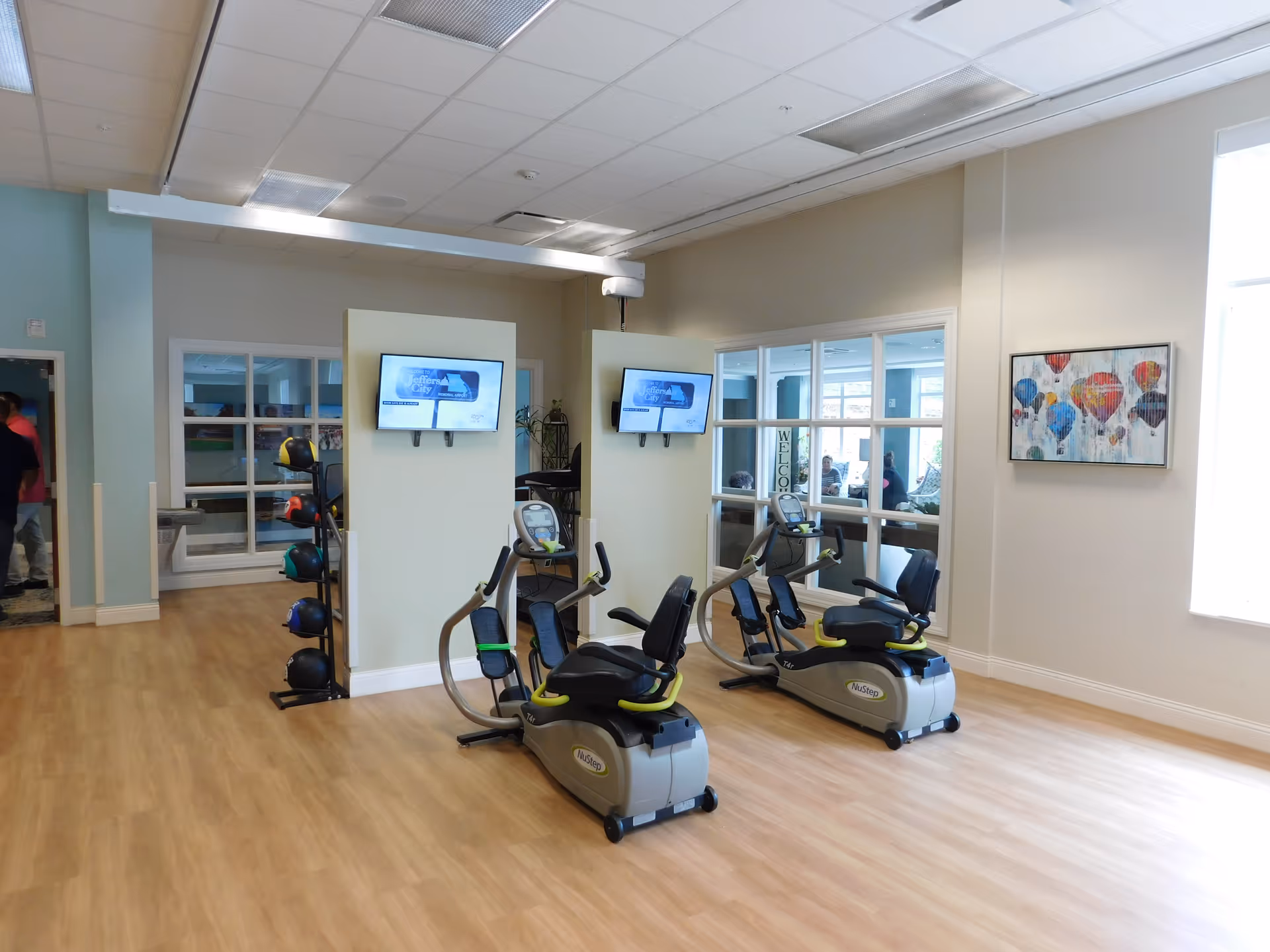 A bright exercise room with two NuStep recumbent cross trainers facing two mounted flat-screen TVs. There is a rack with various medicine balls and a large window with a view into another room where people are seated. The floor is light wood, and the walls are painted in neutral tones with a colorful hot air balloon painting on one wall.