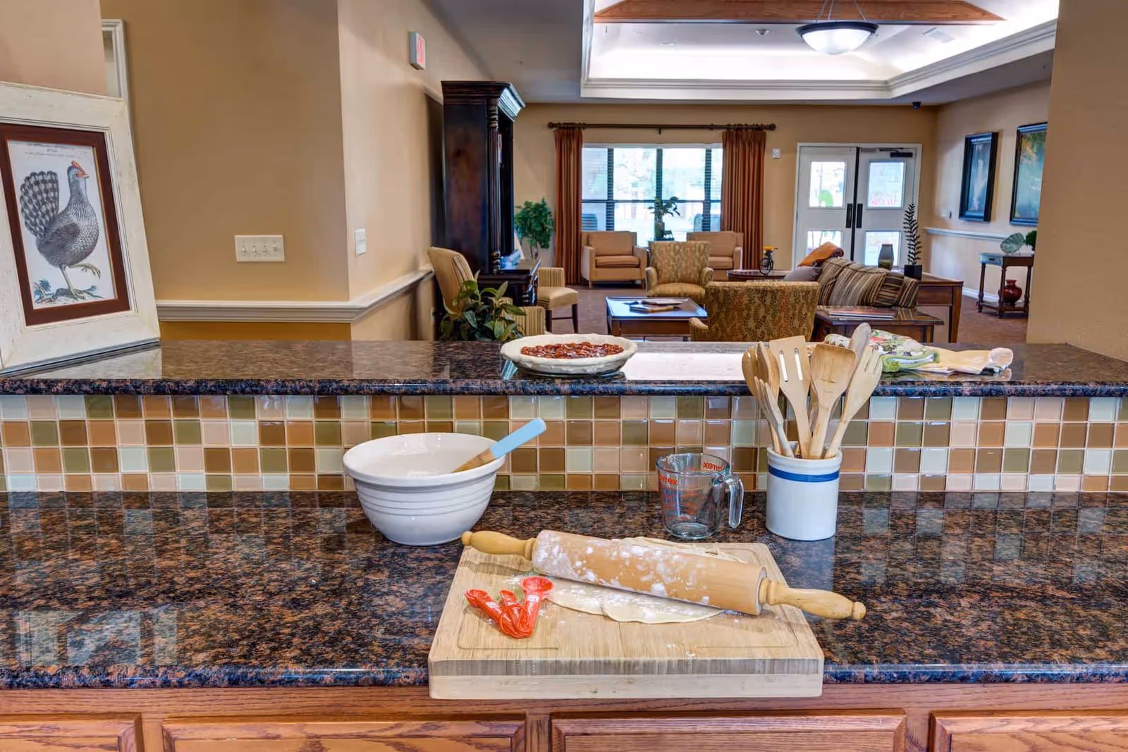 A kitchen counter with baking utensils including a rolling pin, measuring spoons, a mixing bowl with a spatula, and a measuring cup. In the background, there is a living room area with chairs, a sofa, a coffee table, and large windows with curtains.