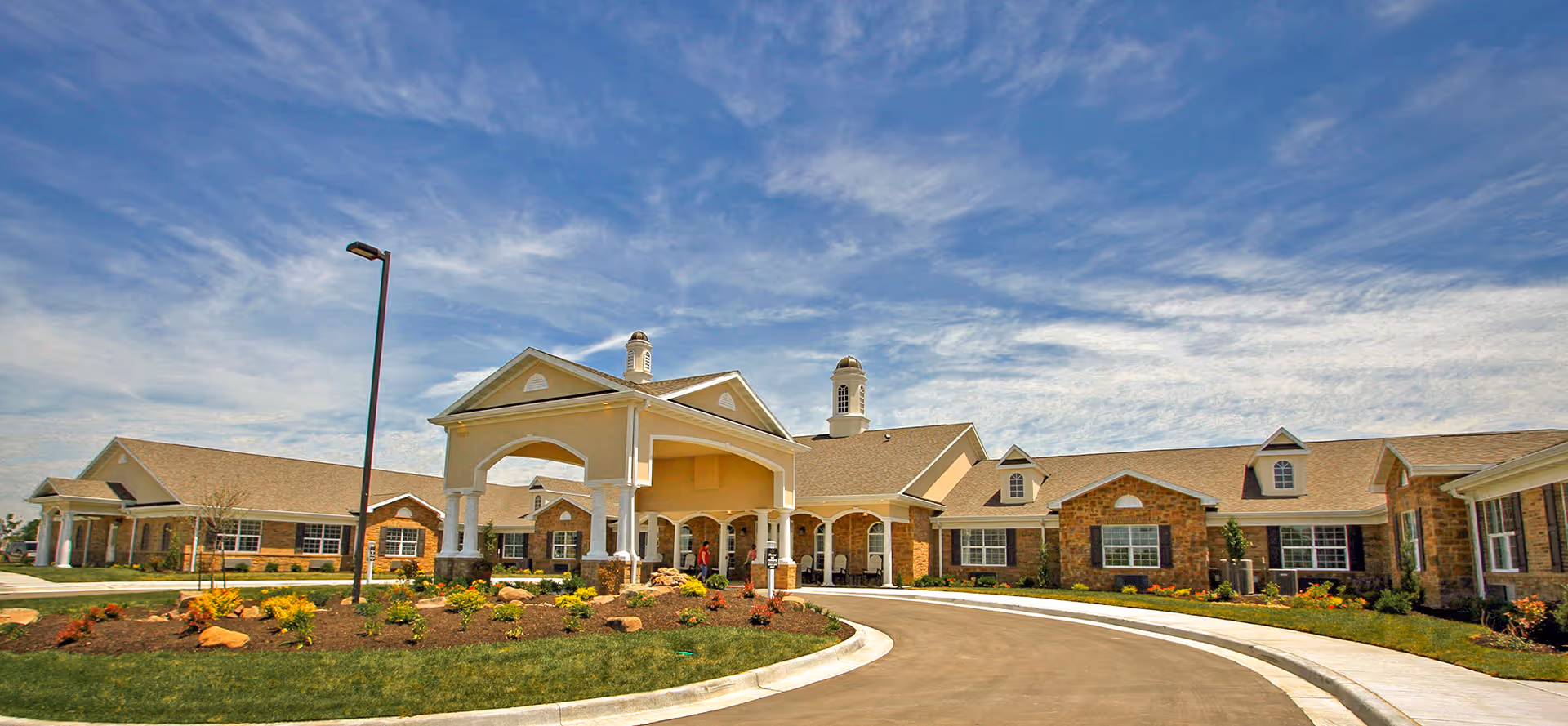 Exterior view of Benton House of Staley Hills, a single-story senior living facility with a covered entrance, landscaped garden beds, and a curved driveway under a partly cloudy blue sky.