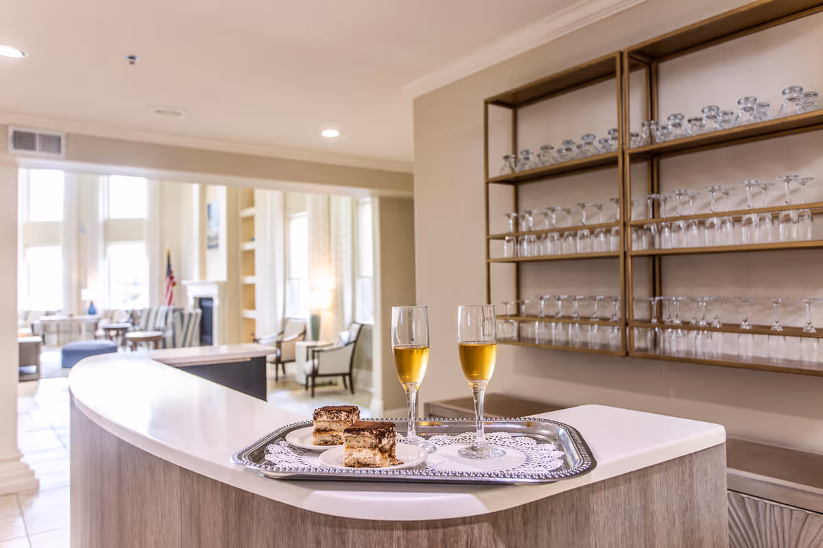 Tray with two glasses of white wine and two dessert slices on a curved counter in a bright lounge with glassware shelves and seating in the background.