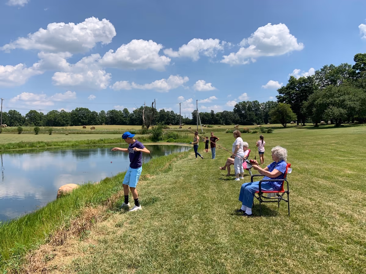 A group of people, including elderly individuals and children, are fishing by a small pond in a grassy outdoor area under a blue sky with scattered clouds. Some are sitting on chairs while others stand near the water's edge, surrounded by open fields and trees in the background.