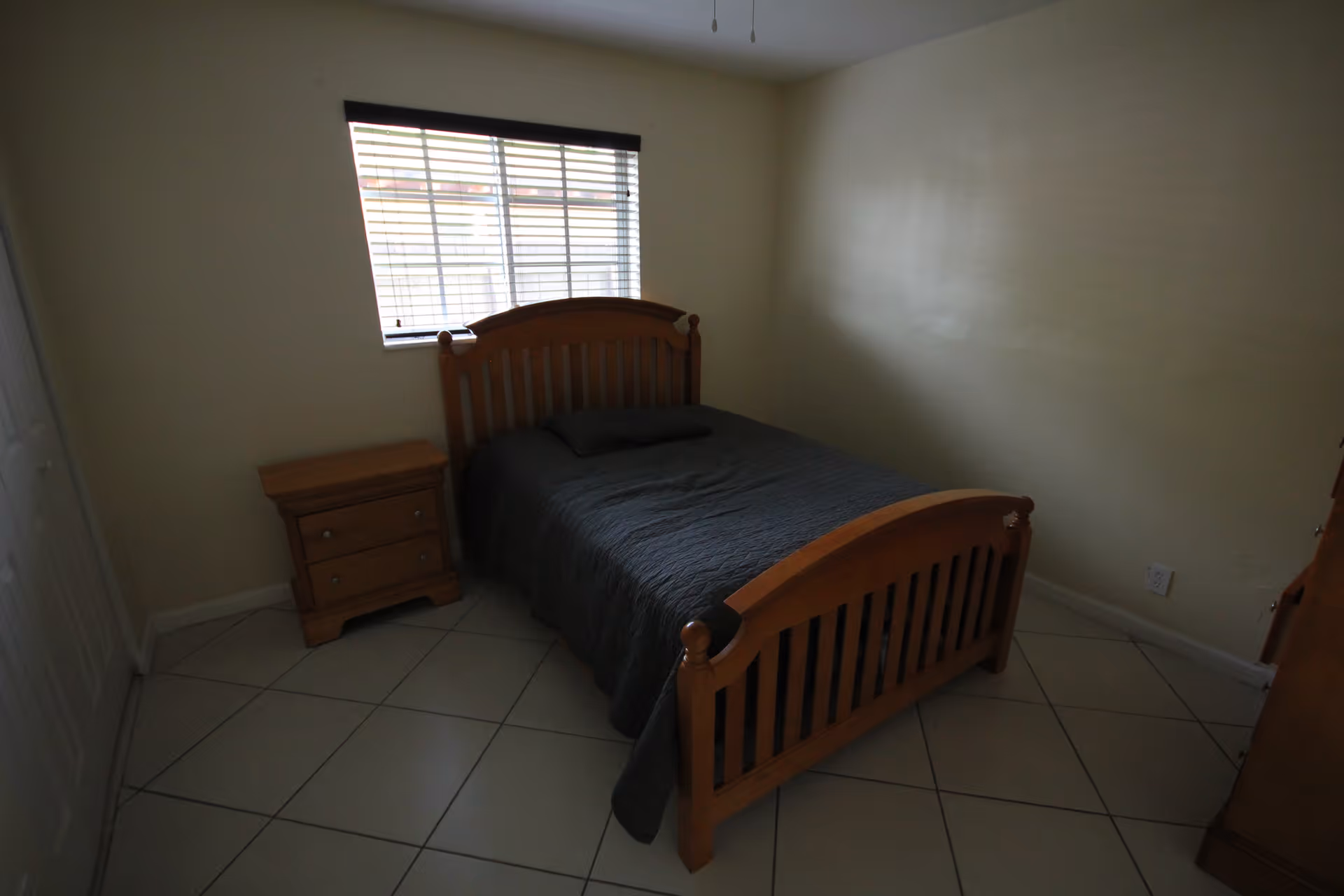 A small bedroom with a wooden bed frame and a dark gray bedspread. There is a wooden nightstand with two drawers next to the bed. The room has tiled flooring and a window with blinds letting in some light. The walls are painted a light beige color.