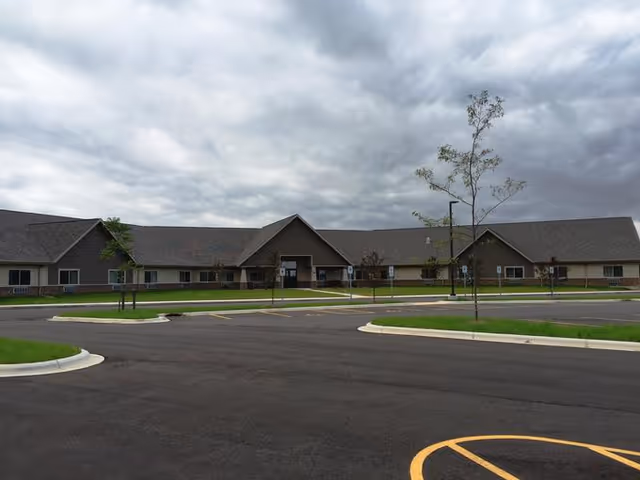 Exterior view of a single-story senior living facility building with a large parking lot in front, a few small trees, and a cloudy sky overhead.
