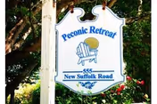 A white hanging sign with blue text and graphics that reads 'Peconic Retreat 555 New Suffolk Road' with an illustration of a chair and sun, surrounded by green foliage and trees.