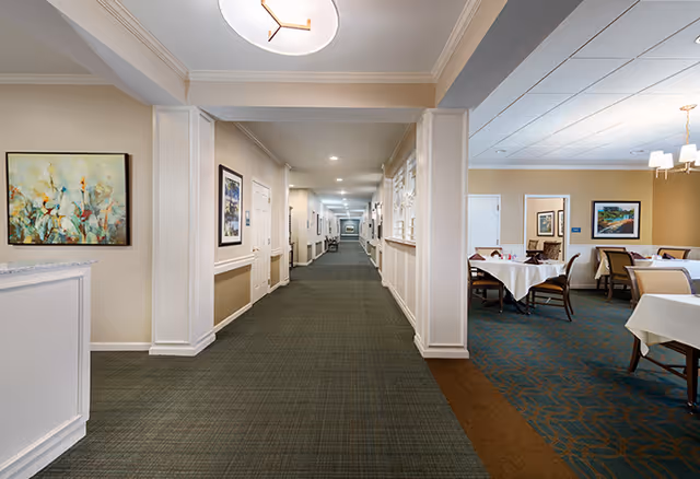 A long, well-lit hallway in a senior living facility with carpeted floors and framed artwork on the walls. To the right, there is a dining area with tables covered in white tablecloths and chairs arranged around them. The ceiling has recessed lighting and a modern circular light fixture is visible in the hallway.