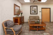 A cozy sitting area in a nursing center featuring a cushioned armchair, a wooden coffee table, a small sofa, and a wooden cabinet with a floral arrangement and framed artwork on the walls.