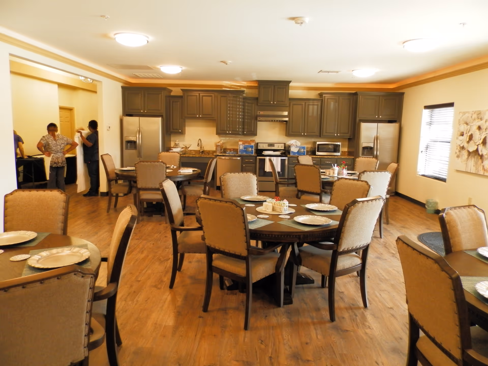 A spacious dining area with multiple round tables set with plates and placemats. The room features wooden flooring and beige upholstered chairs. In the background, there is a kitchen area with gray cabinets, stainless steel appliances including two refrigerators, a stove, a microwave, and a dishwasher. Three people are standing near the entrance to the kitchen area.