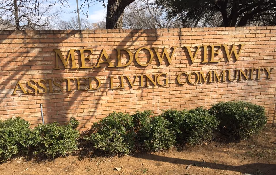 A brick wall with gold letters spelling out 'MEADOW VIEW ASSISTED LIVING COMMUNITY' with green bushes planted in front of the wall and trees visible in the background.