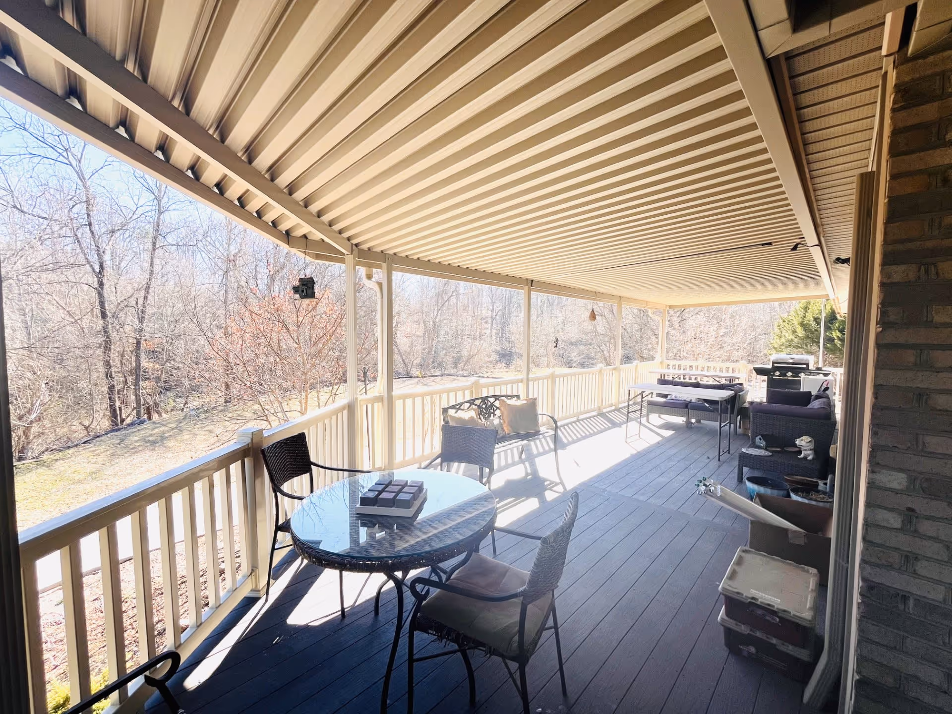 Covered outdoor porch area with a round glass table and four chairs, a bench with cushions, a barbecue grill, and various potted plants and storage containers. The porch overlooks a wooded area with leafless trees.