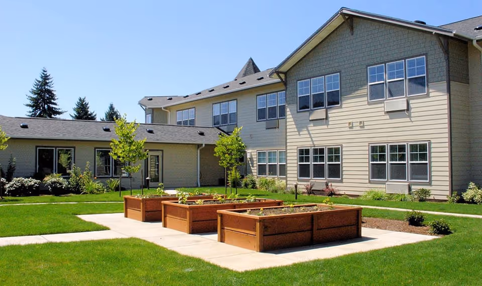 Courtyard with raised wooden garden beds and lawn in front of a two-story beige assisted living building.