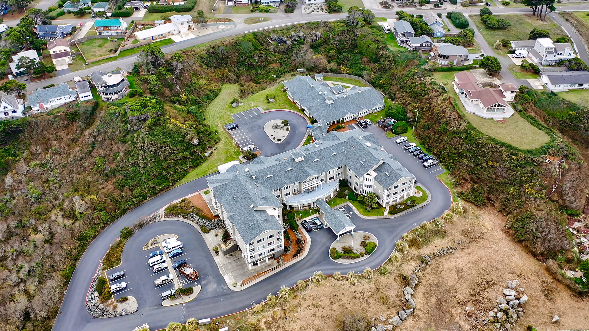 Aerial view of a senior living community complex on a coastal bluff with parking lots, driveways, and nearby houses.