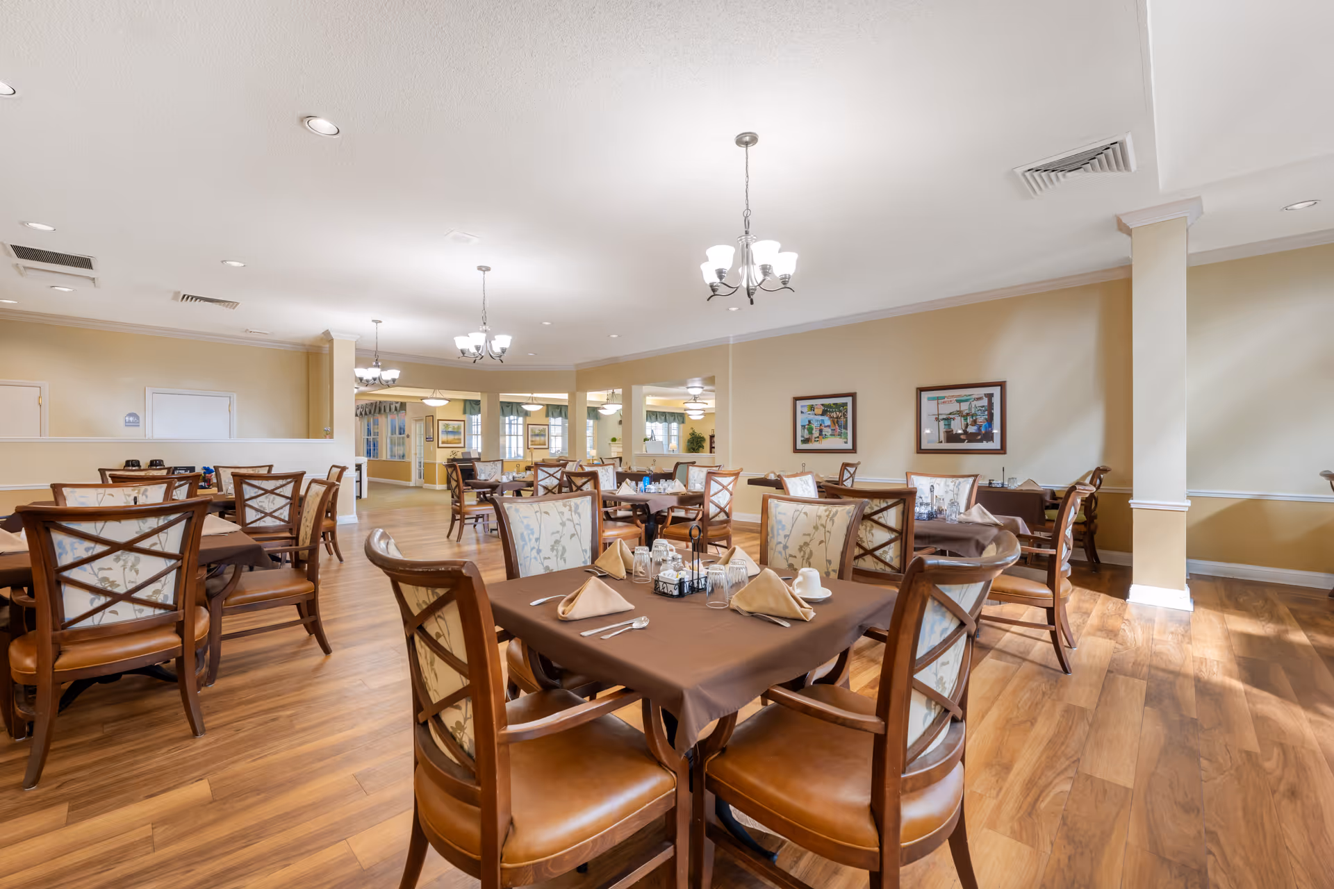 Bright senior living dining room with multiple set tables and wooden chairs on hardwood floors under chandeliers.