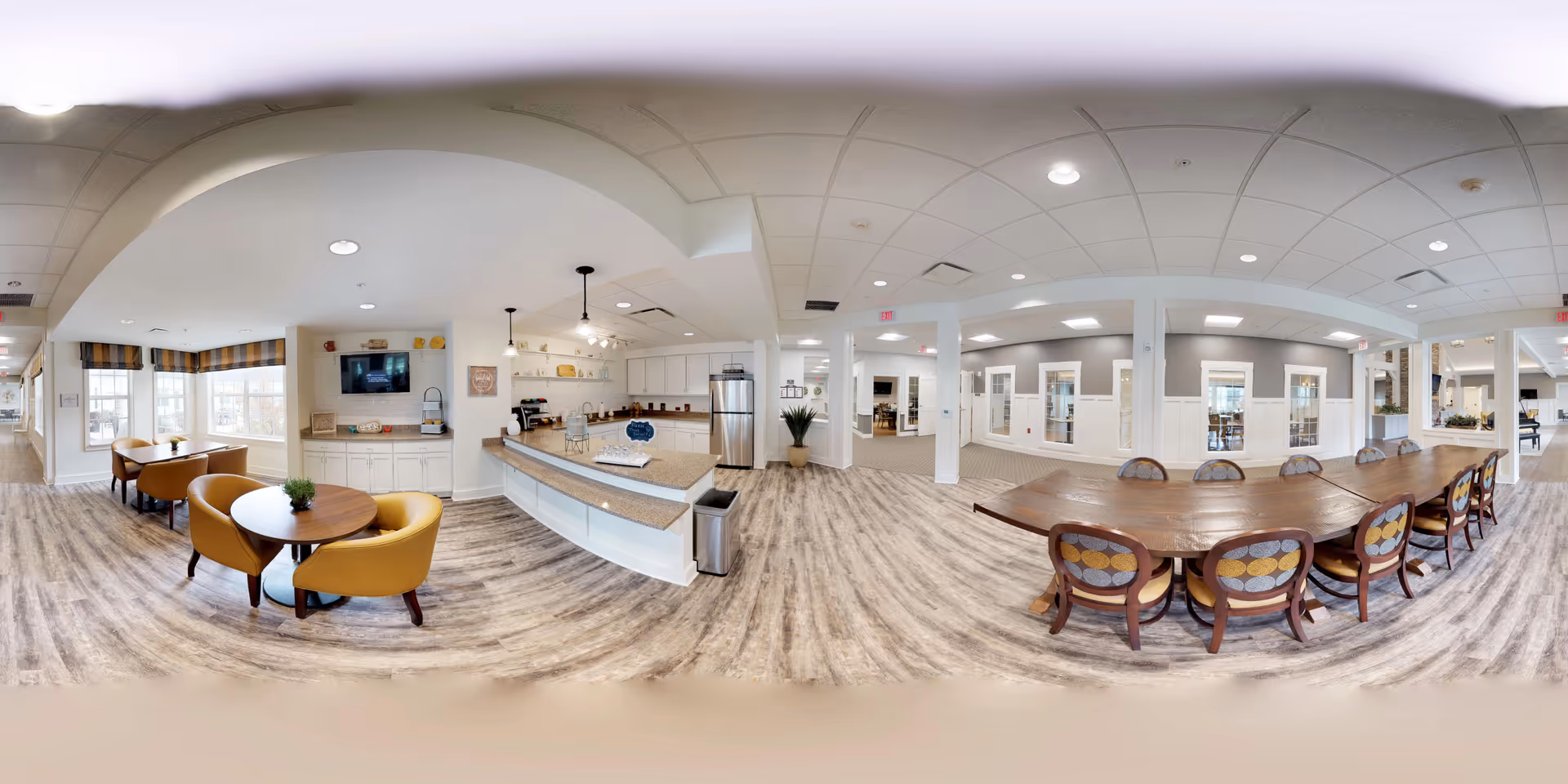 A spacious and well-lit common area in Walnut Crossing featuring a kitchen with a large island, modern appliances, and pendant lighting. To the left, there are small round tables with yellow chairs near large windows with striped valances. To the right, a long wooden table with multiple chairs is set up for group dining or activities. The flooring is light wood, and the ceiling has recessed lighting with a white and neutral color scheme throughout.