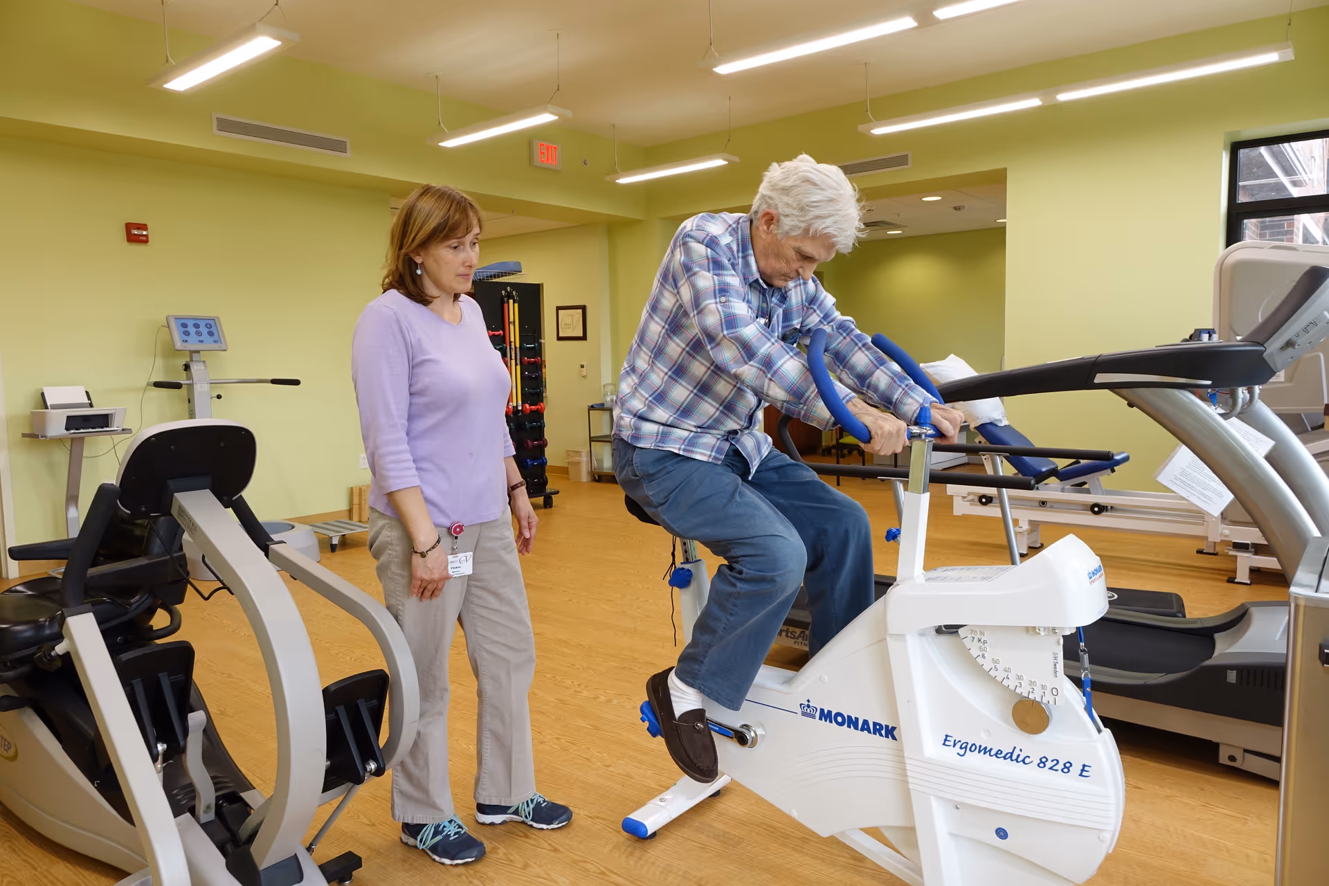 An elderly man is exercising on a stationary bike in a fitness room while a woman, possibly a caregiver or trainer, stands nearby observing him. The room has light green walls, wooden flooring, and various exercise equipment including treadmills and other fitness machines.