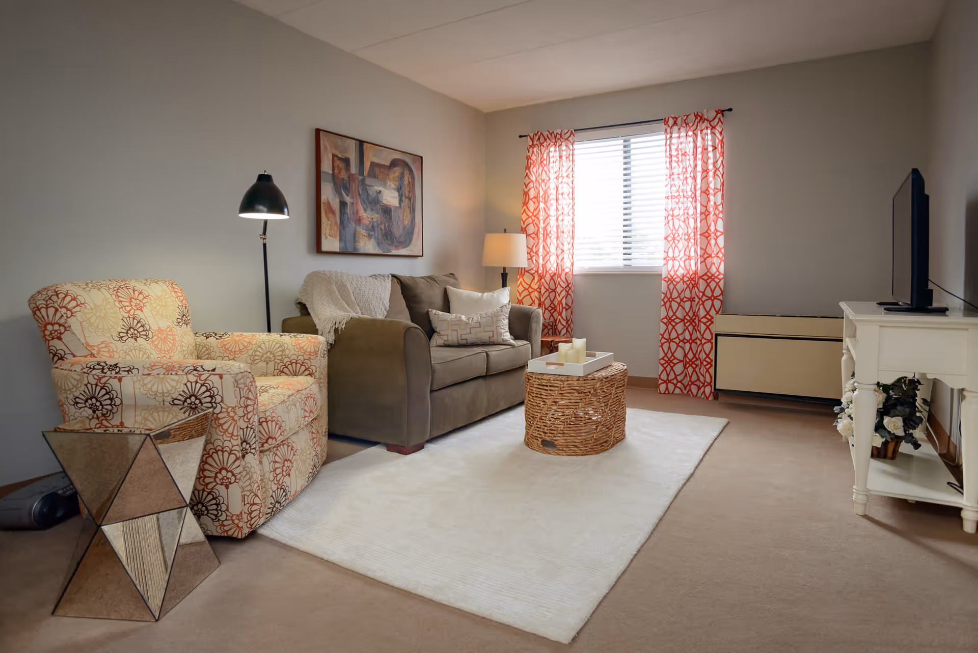 A cozy living room with a patterned armchair, a brown sofa with pillows and a throw blanket, a wicker coffee table with candles on a tray, a white rug, a floor lamp, a table lamp, a window with red patterned curtains, a white TV stand with a flat-screen TV, and a piece of abstract artwork on the wall.