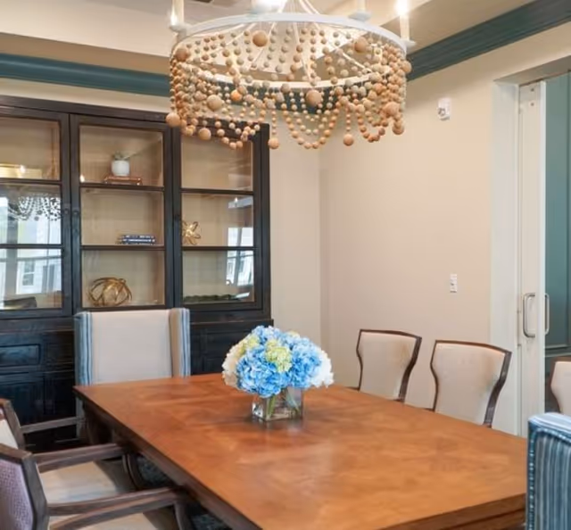 A dining room with a wooden table surrounded by upholstered chairs. A glass vase with blue and white flowers is placed in the center of the table. Behind the table is a dark wooden cabinet with glass doors displaying decorative items. A chandelier with wooden bead decorations hangs above the table.