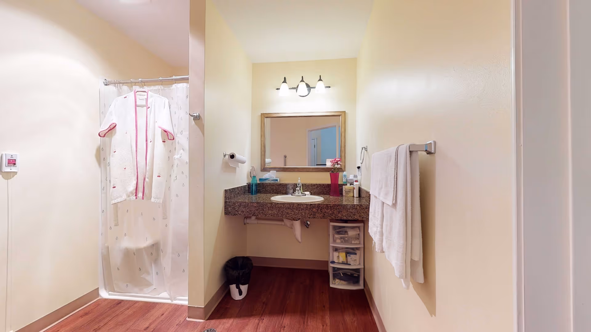 A clean and well-lit bathroom featuring a shower with a white curtain and a white robe hanging on the shower rod. There is a granite countertop with a sink, a mirror above it, and a three-light fixture. A towel rack with a white towel is mounted on the wall to the right, and a small plastic drawer unit is placed under the counter. The floor has a wood-like finish.