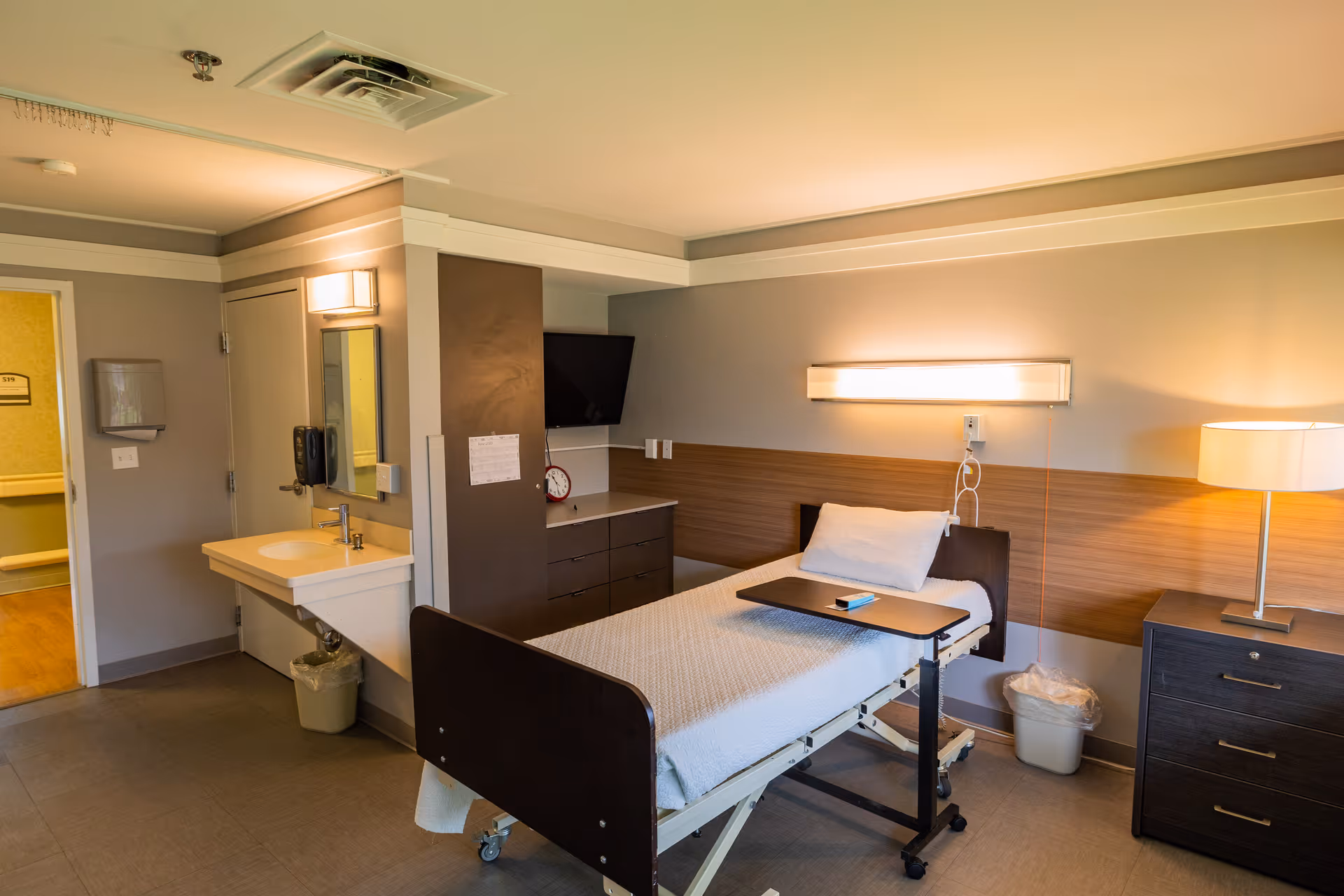 A well-lit nursing facility patient bedroom with a hospital bed, overbed table, sink, dresser, TV, and bedside lamp.