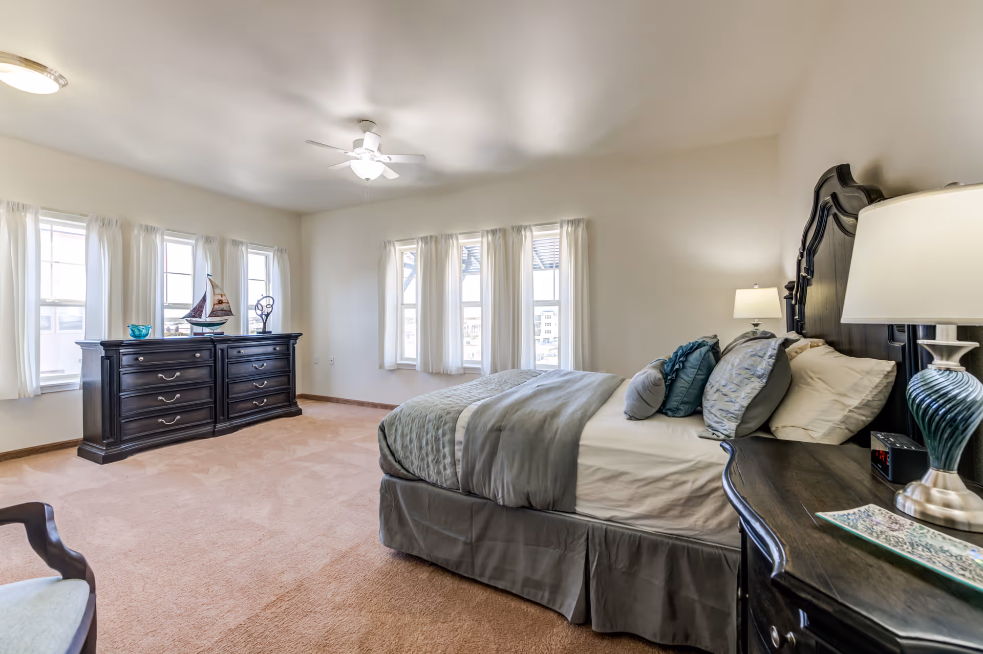 A spacious bedroom with beige carpet and cream-colored walls. The room features a large bed with gray and blue bedding and multiple pillows. On the right side of the bed is a dark wooden nightstand with a lamp and a small clock. Across from the bed is a matching dark wooden dresser with decorative items including a model sailboat and a sculpture. Several windows with white curtains allow natural light to fill the room. A ceiling fan with a light fixture is mounted on the ceiling.