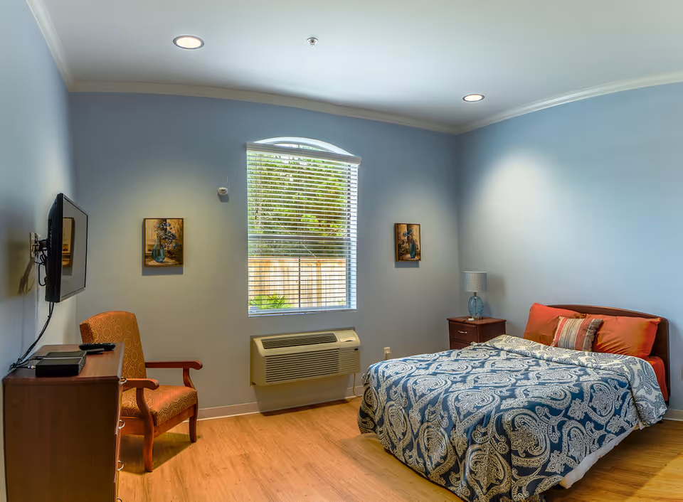 A cozy bedroom with light blue walls, a window with blinds, a bed with a blue and white patterned comforter and red pillows, a wooden nightstand with a lamp, a wooden dresser with a mounted flat-screen TV above it, and an upholstered armchair with wooden arms.