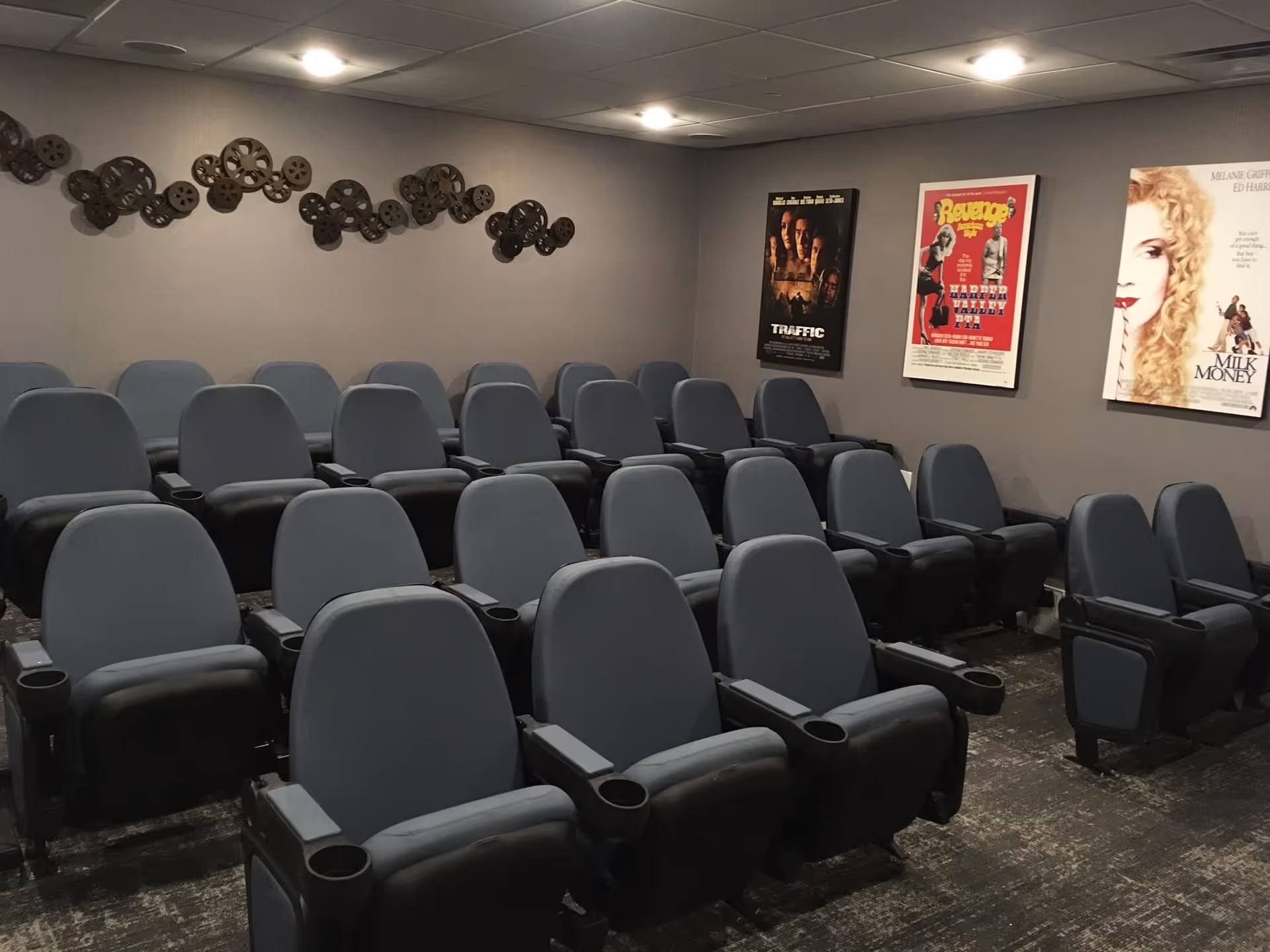 A small theater room with several rows of gray cushioned seats with armrests and cup holders. The walls are gray and decorated with movie posters and a metal wall art piece resembling film reels.