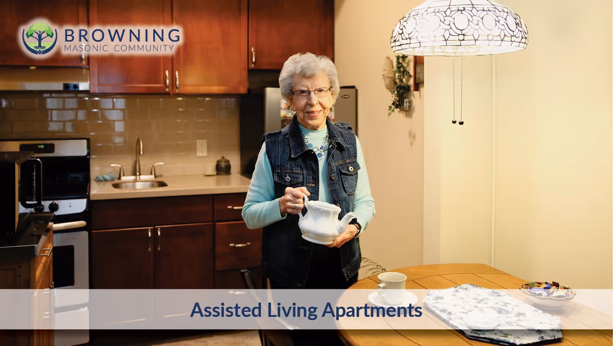 An elderly woman standing in a kitchen holding a white teapot. The kitchen has wooden cabinets, a sink, a stove, and a refrigerator. A round wooden table with a cup and saucer, a bowl of candy, and a floral placemat is visible. A decorative hanging lamp is above the table. The image includes the Browning Masonic Community logo and the text 'Assisted Living Apartments'.