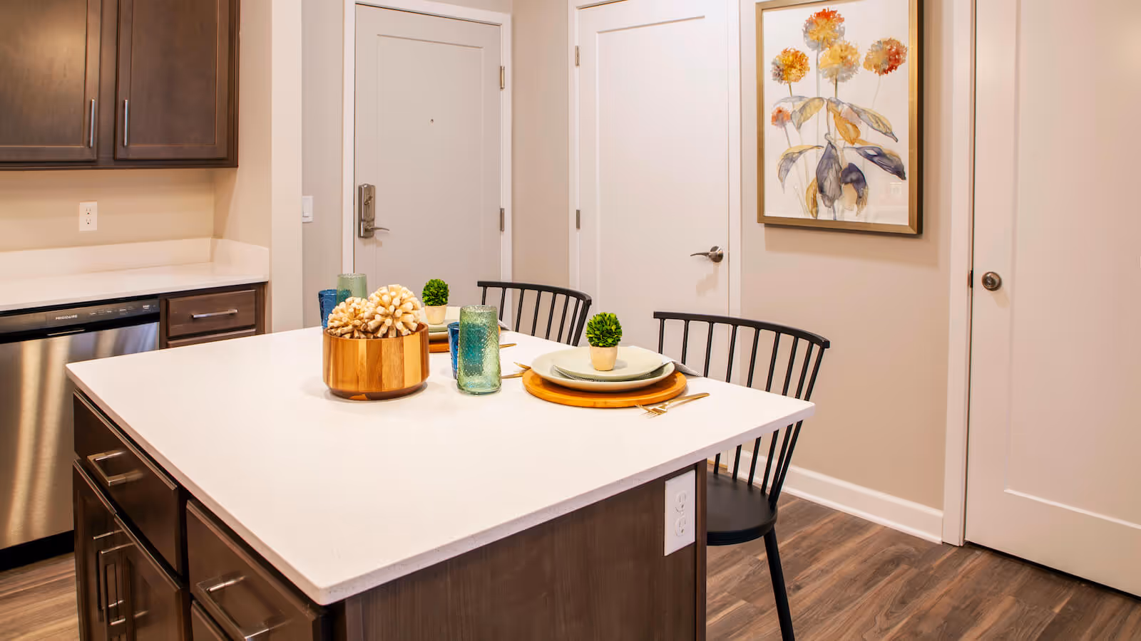 Bright kitchen interior with a white island set with plates and chairs, dark cabinets, and framed artwork on the wall.