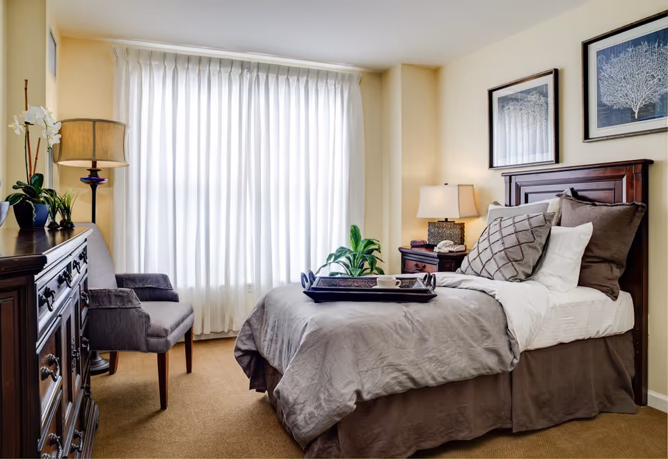 A cozy bedroom with a neatly made bed featuring multiple pillows and a tray with a cup on it. There is a wooden nightstand with a lamp and a telephone, a wooden dresser with plants and a lamp, and a gray upholstered chair. The room has beige walls, carpeted floor, and two framed artworks above the bed. A large window with white curtains allows natural light to fill the room.