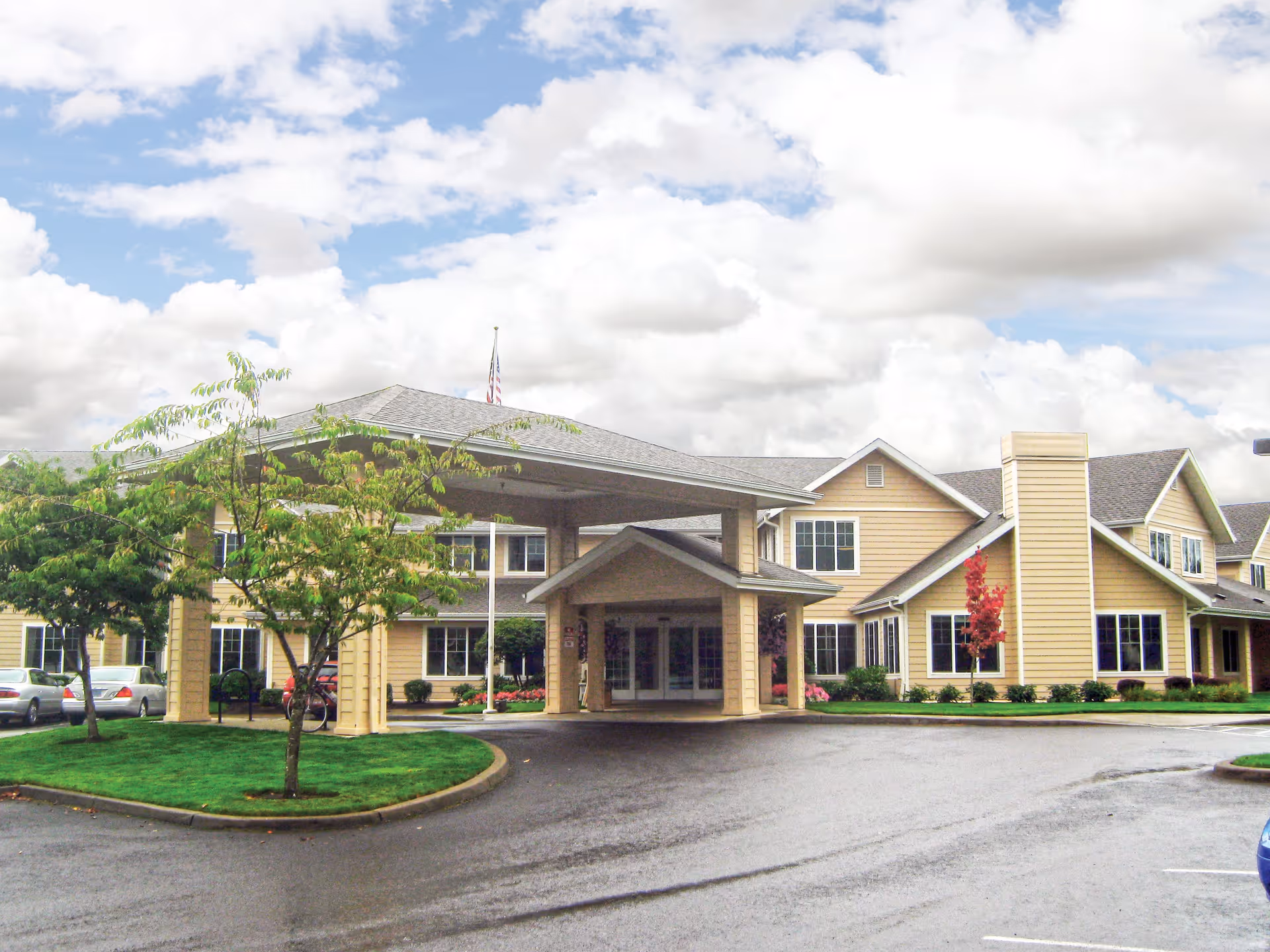 Exterior view of a senior living facility named Stoneybrook Senior Living with a covered entrance, beige siding, multiple windows, a well-maintained lawn, and trees under a partly cloudy sky.